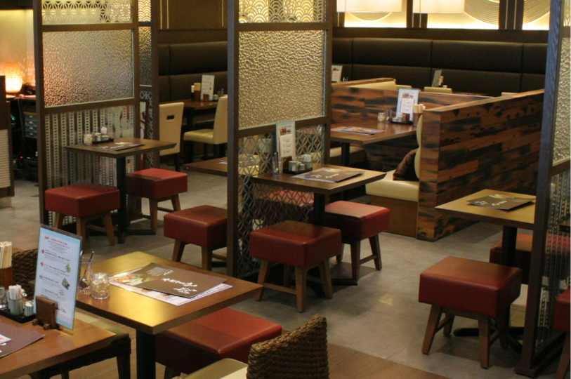 Interior of a cozy Japanese restaurant featuring warm wood tables and red cushioned stools. Decorative frosted glass partitions with geometric patterns separate the dining areas, and a rustic wood-paneled booth is visible in the background.