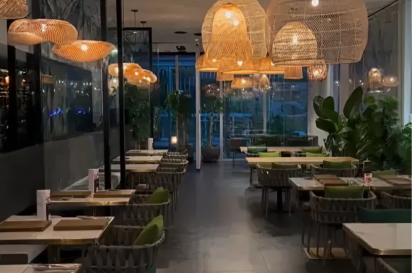A modern, dimly lit restaurant interior characterized by numerous large, woven wicker pendant lights hanging at various heights. The space includes sleek white tables, grey woven chairs with green cushions, and large indoor plants, creating an upscale, organic atmosphere.