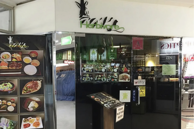 The storefront of "Herbivore," a Japanese vegetarian restaurant. The entrance features a large glass door and a white wall above it with a stylized zebra logo. A menu stand and posters showcasing various colorful bento boxes and sushi platters are displayed prominently at the entrance.