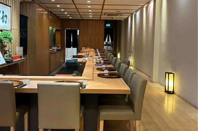 A minimalist and elegant Japanese omakase dining room. A long, light-colored wooden L-shaped counter with grey leather chairs dominates the room. The design is clean, featuring warm wood paneling, soft recessed ceiling lights, and glowing floor lanterns along a textured white wall.
