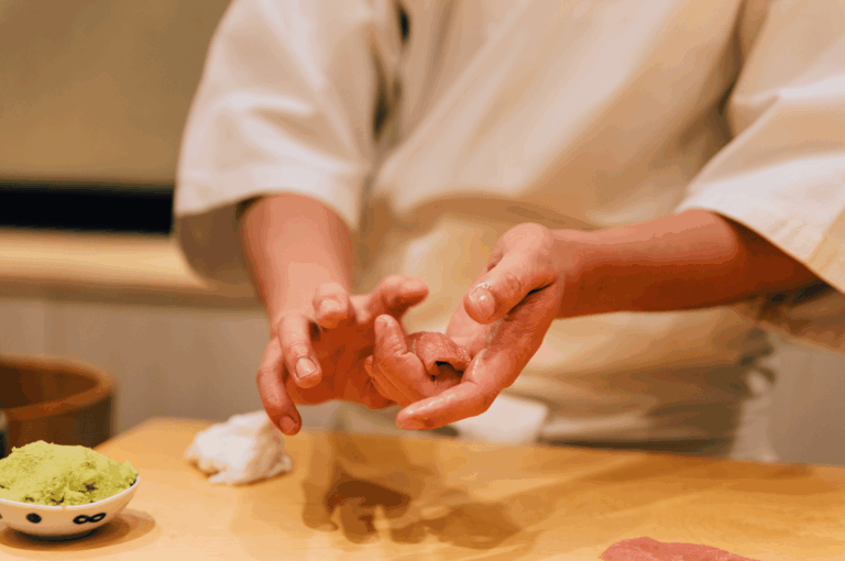 A sushi chef in a crisp white uniform delicately molds a piece of nigiri, capturing the quiet precision and reverence of omakase preparation. His hands, mid-motion, reflect years of mastery, while a small bowl of wasabi and a neatly folded cloth suggest readiness and ritual. The intimate setting evokes the beginning of a culinary journey where every gesture is part of the welcome.