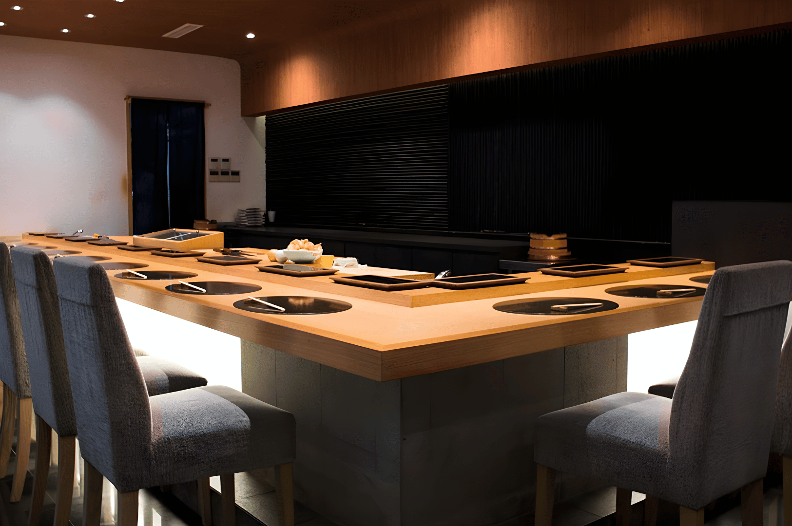 A softly lit U-shaped sushi counter awaits its next guest, each place setting meticulously arranged with chopsticks, plates, and round placemats. The warm glow from beneath the counter and the sleek black slatted wall evoke a serene, modern intimacy. This final scene captures the farewell moment of omakase—where the space itself bows in gratitude, leaving behind the memory of a shared culinary ritual.