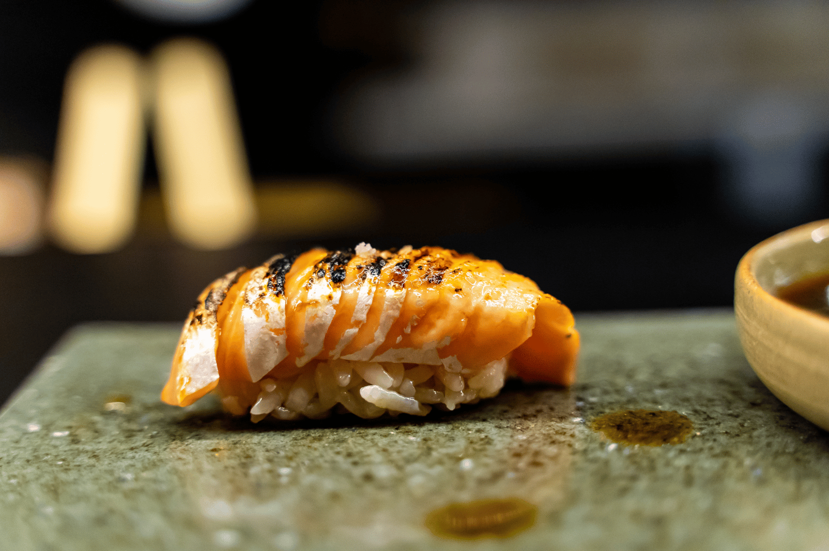 A seared salmon nigiri rests atop a green ceramic surface, its charred grill marks glistening with droplets of sauce. The subtle blur of the background draws focus to the interplay of texture and color—smoky orange against earthy green. This moment in the omakase sequence evokes comfort and depth, a gentle crescendo of flavor and intimacy before the evening’s quiet descent.