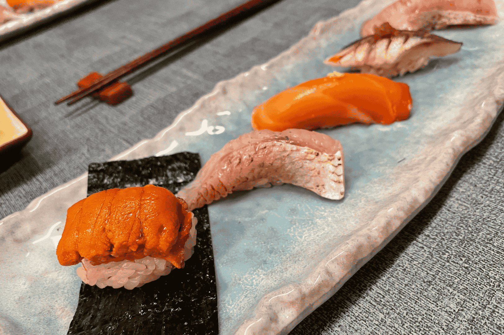 Assorted nigiri and maki sushi arranged on a white rectangular plate with chopsticks, shot from above on a neutral background—clean, contemporary presentation.