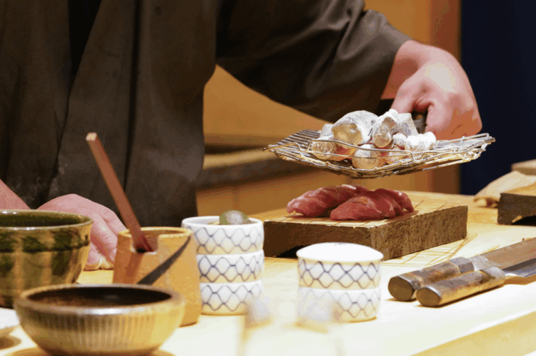 A sushi chef at Ginza Sushi Ichi sears ingredients over hot charcoal at a traditional wooden counter, highlighting artisanal technique and premium presentation. Two pieces of nigiri rest on a minimalist wooden board, surrounded by ceramic cups and precision knives—evoking the intimate, high-end ambiance central to the restaurant’s value proposition.