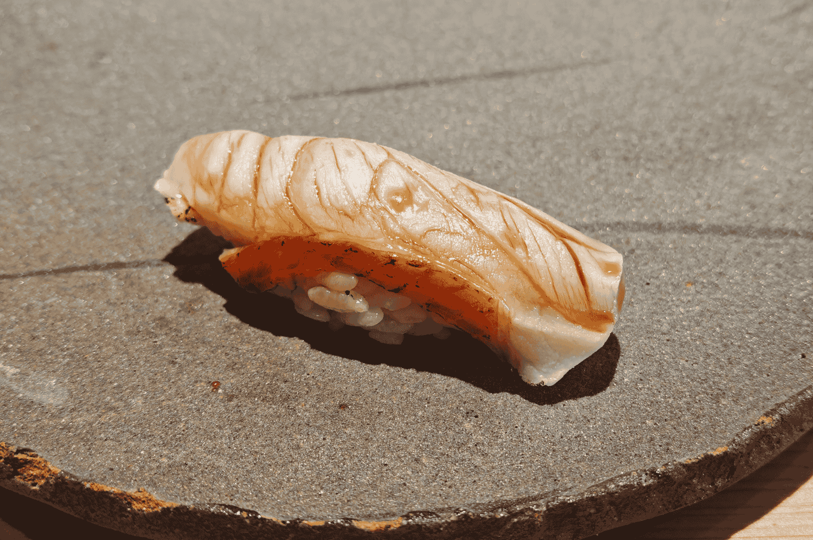 A single piece of aburi-style nigiri at Shinji, presented on a textured ceramic plate. The lightly torched fish glistens with grill marks, emphasizing precision and restraint in flavor layering. The minimalist plating and stoneware surface evoke understated luxury—contrasting Ginza Sushi Ichi’s theatrical craftsmanship with Shinji’s quiet refinement.