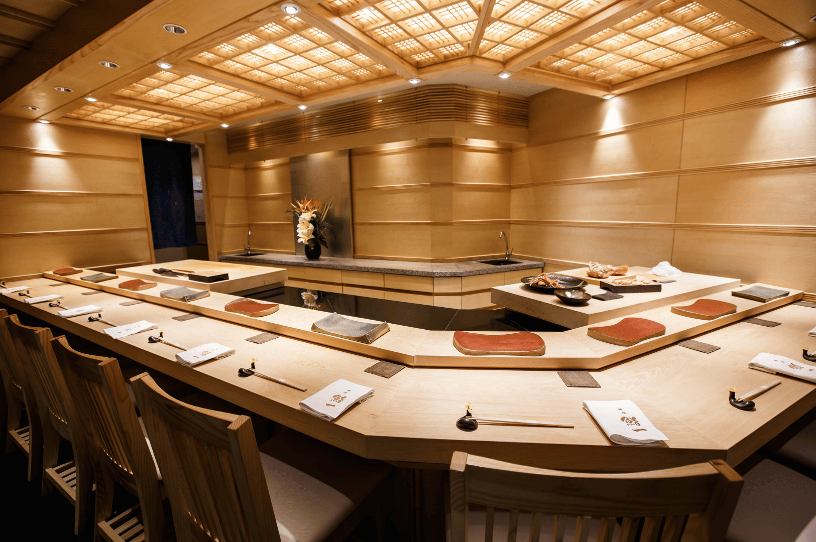 Interior of Ginza Sushi Ichi’s omakase counter, featuring a U-shaped light wood bar with individual place settings and slatted chairs. The warm lighting and grid-patterned ceiling create an intimate yet refined atmosphere, while the decorative mask and floral centerpiece behind the counter hint at theatrical elegance—reinforcing the restaurant’s premium positioning in the value-for-money debate.