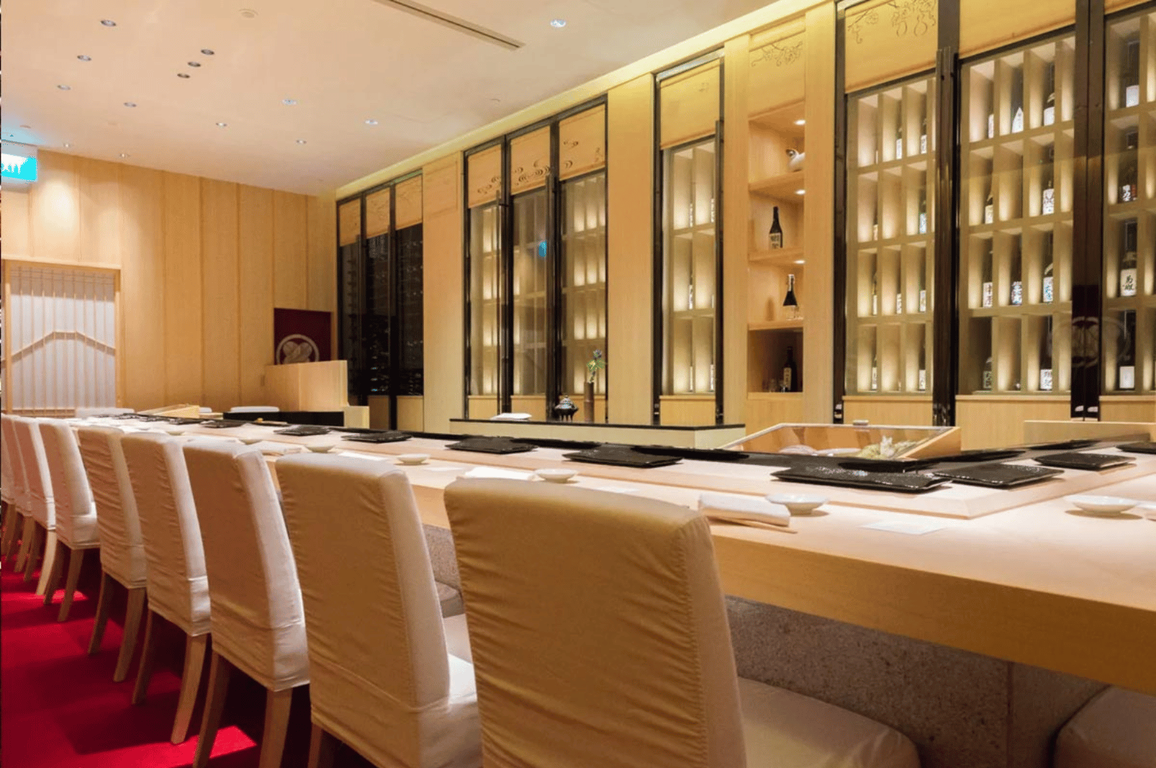 Shinji’s serene sushi counter features a long wooden bar with crisp white fabric-covered chairs and minimalist black place settings. Illuminated shelves in the background display curated bottles and decor, adding quiet sophistication. The soft lighting and restrained design reflect Shinji’s philosophy of elegance through simplicity—offering a subtler, contemplative counterpoint to Ginza Sushi Ichi’s theatrical flair.