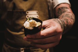 Close-up of a chef’s tattooed hand presenting a gunkan-maki sushi, wrapped in nori and topped with creamy uni and thin cucumber slices. The chef wears a brown apron with white buttons, evoking a warm, artisanal omakase setting. The image captures the intimacy and craftsmanship of Japanese dining, ideal for group omakase experiences in Singapore.