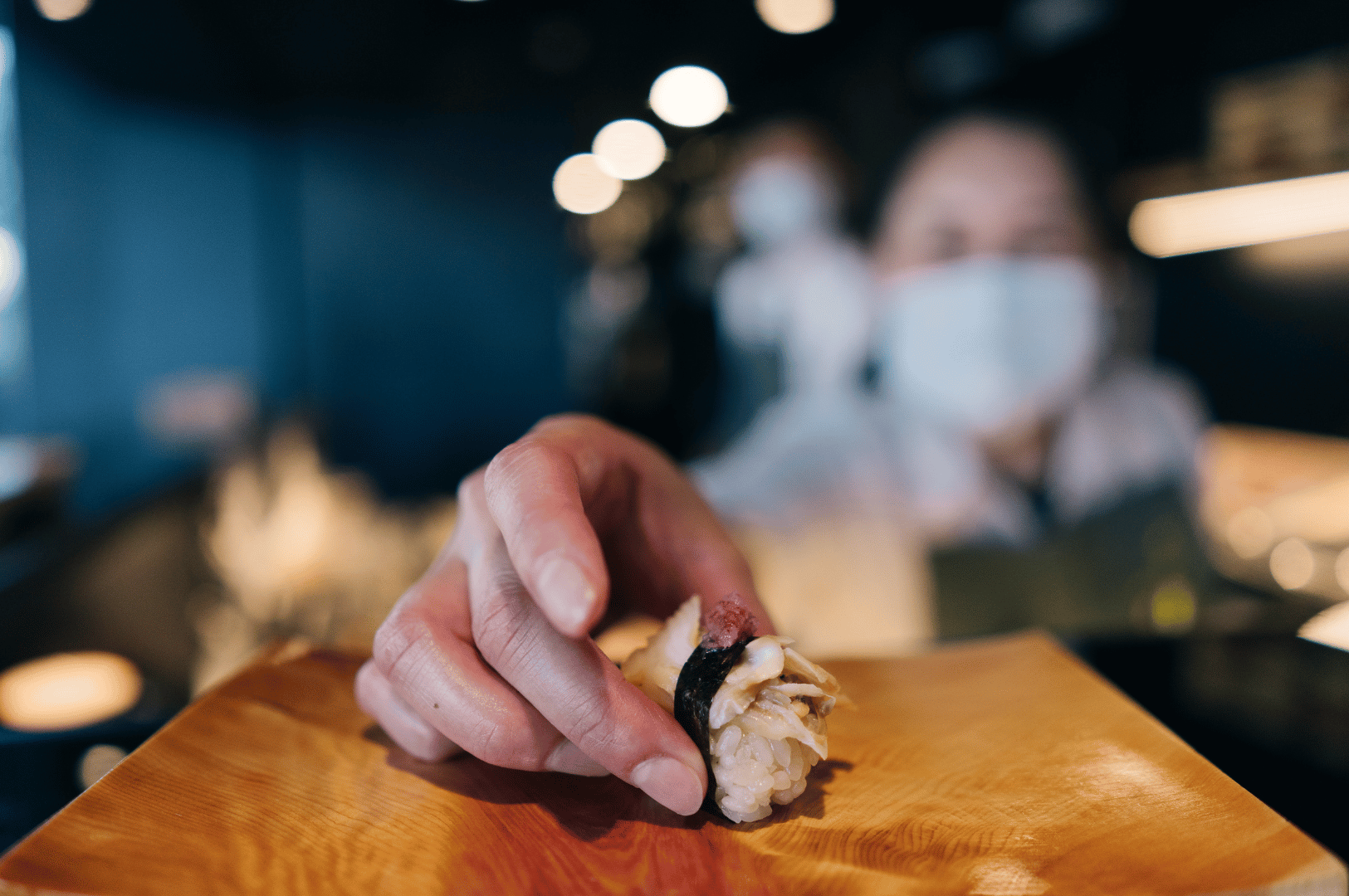 Masked chef in crisp white attire delicately places a shellfish-topped sushi onto a wooden counter, framed by warm bokeh lights and blurred diners in the background. The gesture evokes precision and hospitality, capturing the quiet theater of group omakase dining in Singapore’s top Japanese venues.