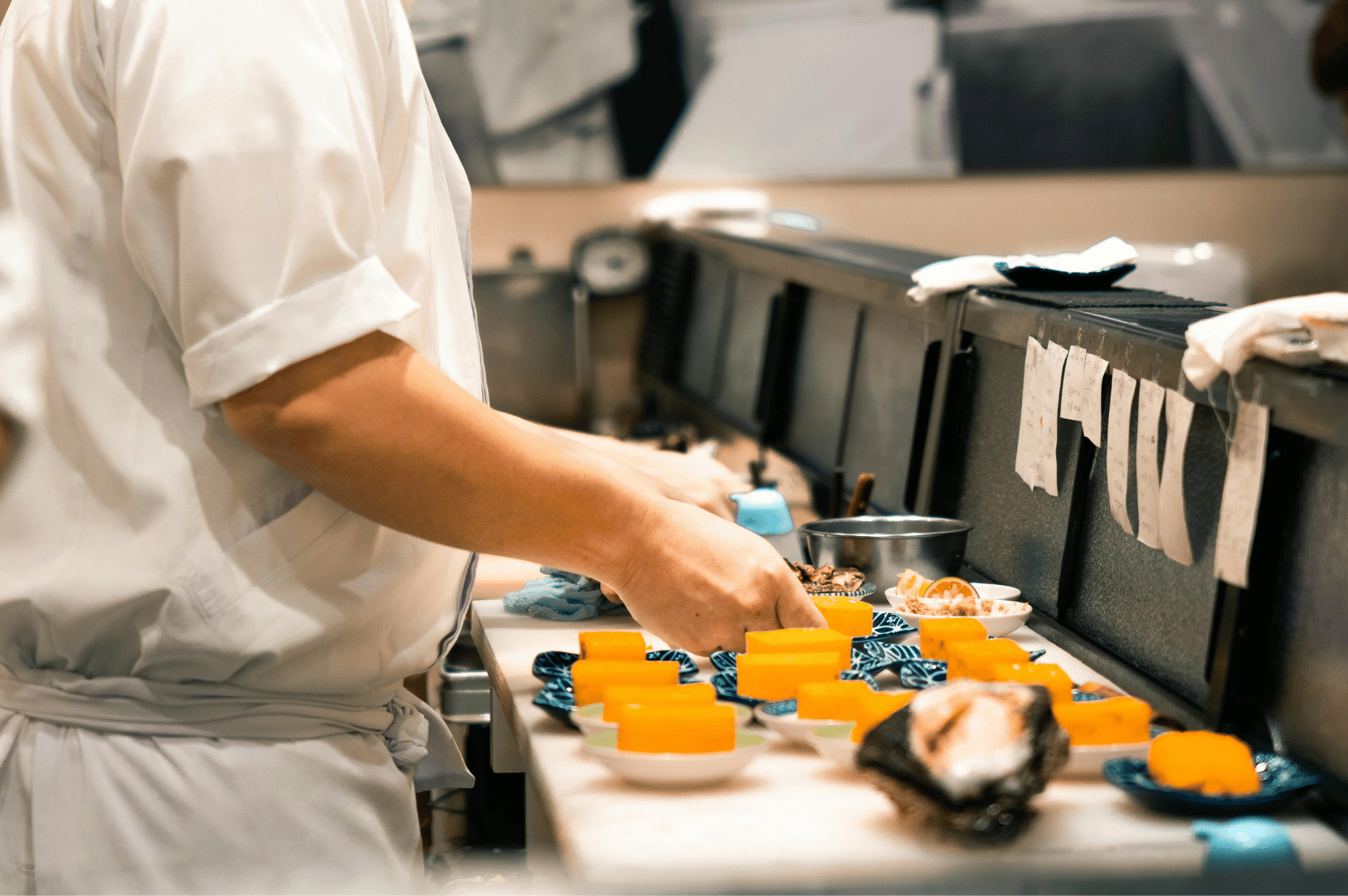 Chef in white uniform prepares vibrant orange tamago on blue ceramic plates, surrounded by oyster shells, bowls, and utensils on a bustling kitchen counter. The scene captures the behind-the-scenes rhythm of group omakase service, where precision and artistry converge in Singapore’s top Japanese dining venues.