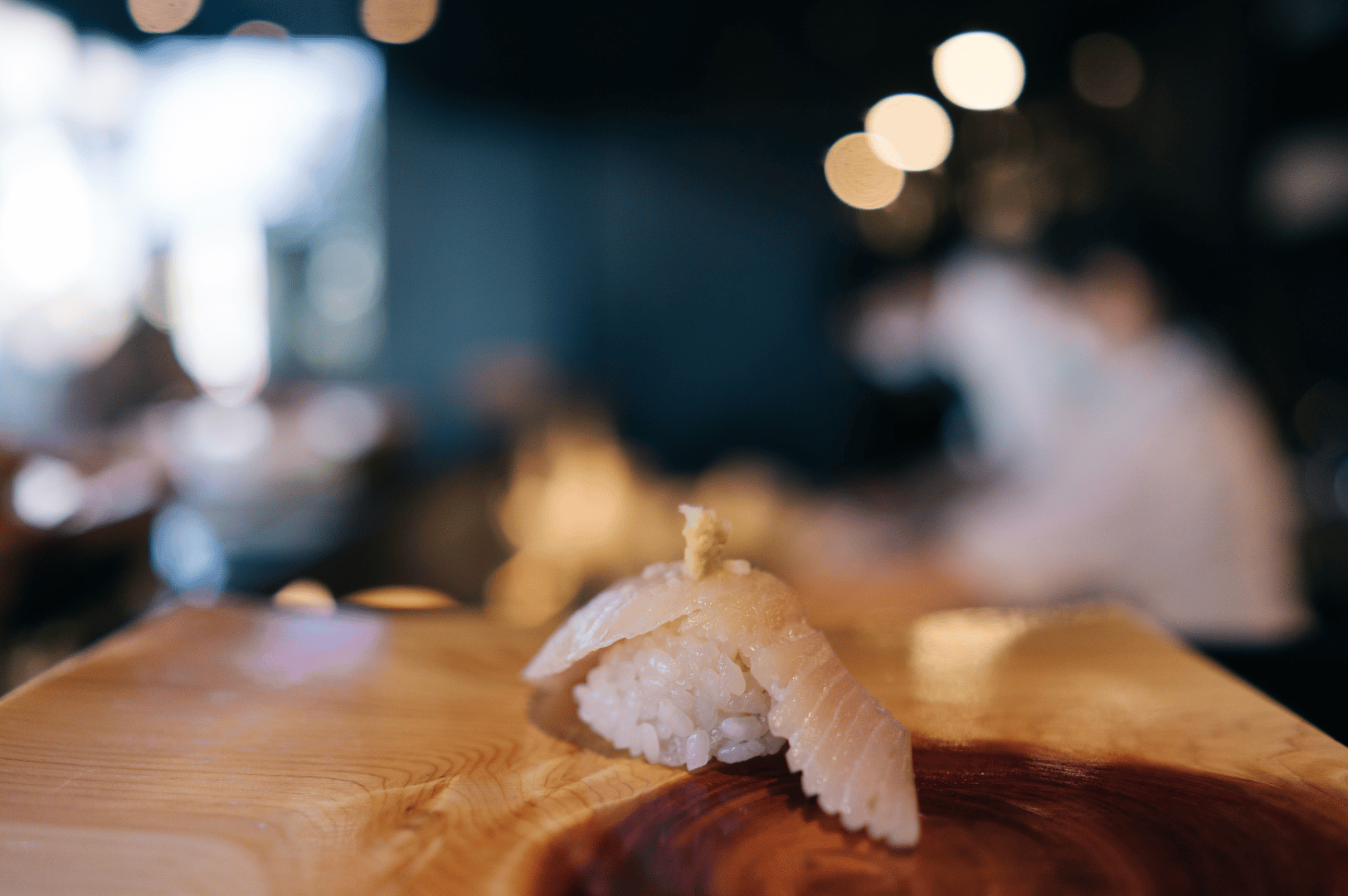 Single piece of white fish sushi garnished with a delicate topping, resting on a wooden counter under soft ambient lighting. Blurred figures and warm bokeh in the background evoke the serene intimacy of upscale group omakase dining in Singapore, where each bite is a moment of crafted stillness.