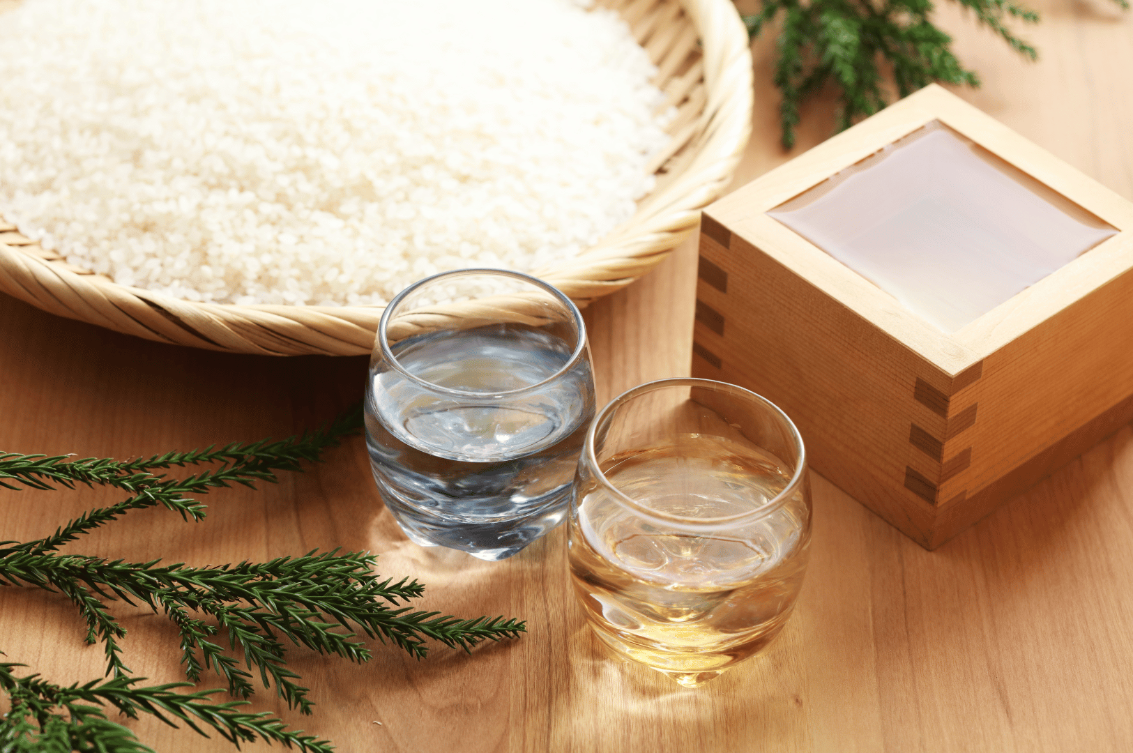 Traditional sake elements arranged with ritual elegance—uncooked white rice in a woven basket, clear glasses of water and sake, and a wooden masu box framed by pine branches. The composition evokes seasonal celebration and craftsmanship, enriching the sensory tapestry of group omakase dining in Singapore’s finest Japanese venues.