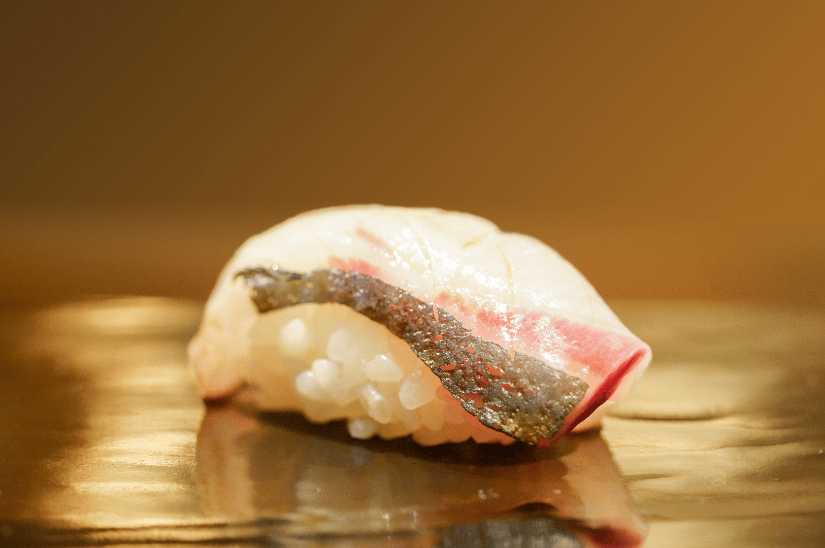 Silver-skinned mackerel sushi glistens atop vinegared rice on a reflective surface, set against a warm, softly blurred background. The translucent sheen and precise cut highlight the purity and elegance of Japanese culinary technique—an understated yet powerful detail in Singapore’s best group omakase venues.