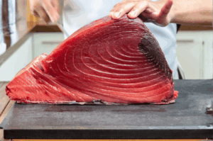 Close-up of a chef preparing a pristine cut of deep red tuna on a wooden board, highlighting the fish’s striated muscle texture and vibrant freshness. The chef’s hands—one steadying the fish, the other wielding a slicing knife—suggest precision and reverence, evoking the meticulous sourcing and butchery rituals behind top omakase menus. Natural lighting and shallow depth of field emphasize the tuna’s quality, framing it as a hero ingredient in the culinary storytelling of elite Japanese dining.