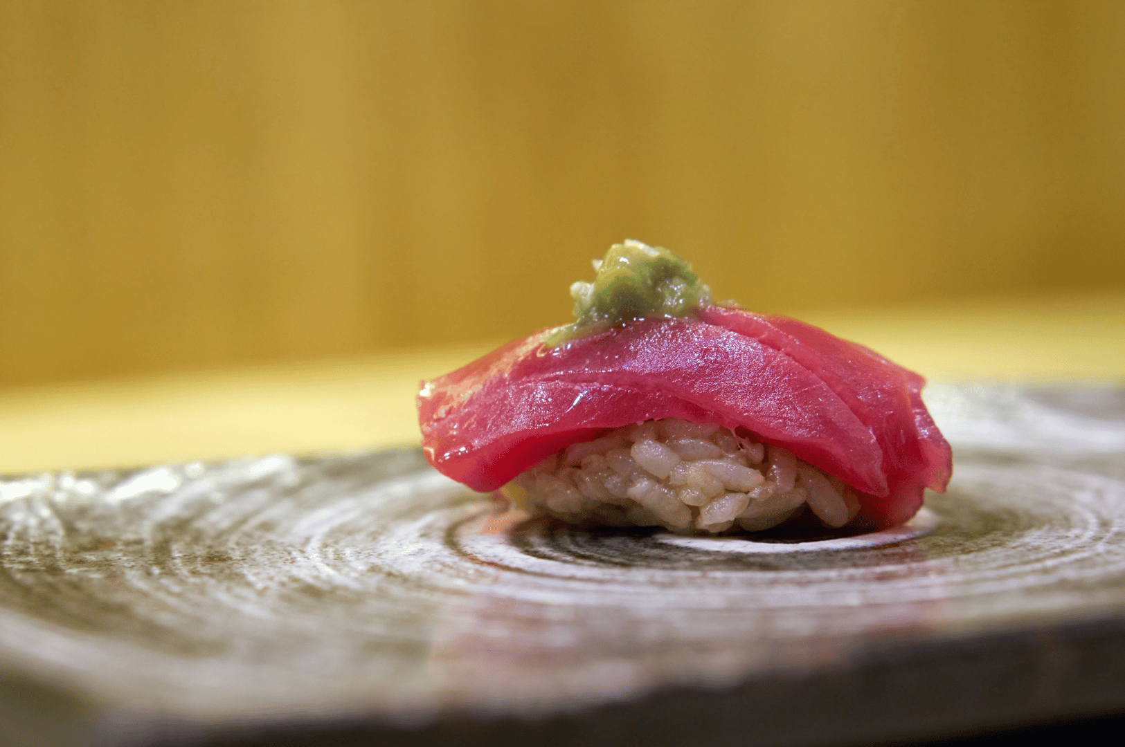 Elegant piece of tuna nigiri sushi resting on a warm-toned wooden surface, with a delicate smear of wasabi atop the glistening fish. The softly blurred background and golden lighting evoke the intimacy of a high-end omakase counter, spotlighting the craftsmanship and ingredient purity that define elite sourcing. The composition celebrates restraint and refinement, capturing the moment where sourcing meets artistry in Japanese culinary tradition.