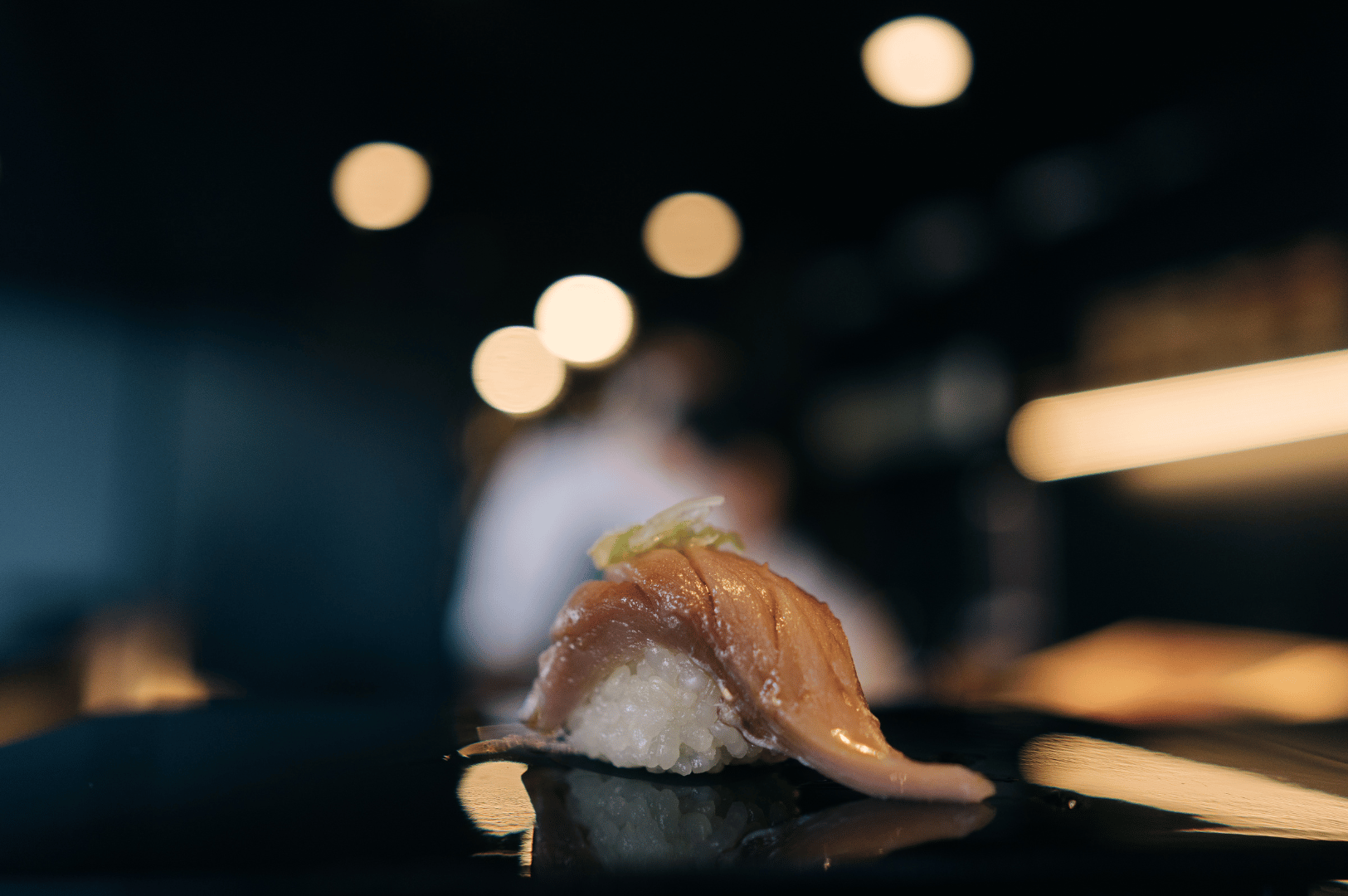 Single piece of sushi—tuna or salmon—presented on a dark surface with warm, ambient lighting and blurred figures in the background, suggesting the presence of chefs or diners. The shallow depth of field draws focus to the glistening fish and delicate rice, capturing the moment of transition from sourcing to service. The composition evokes intimacy and anticipation, reflecting the reverence with which top omakase restaurants present their finest ingredients.