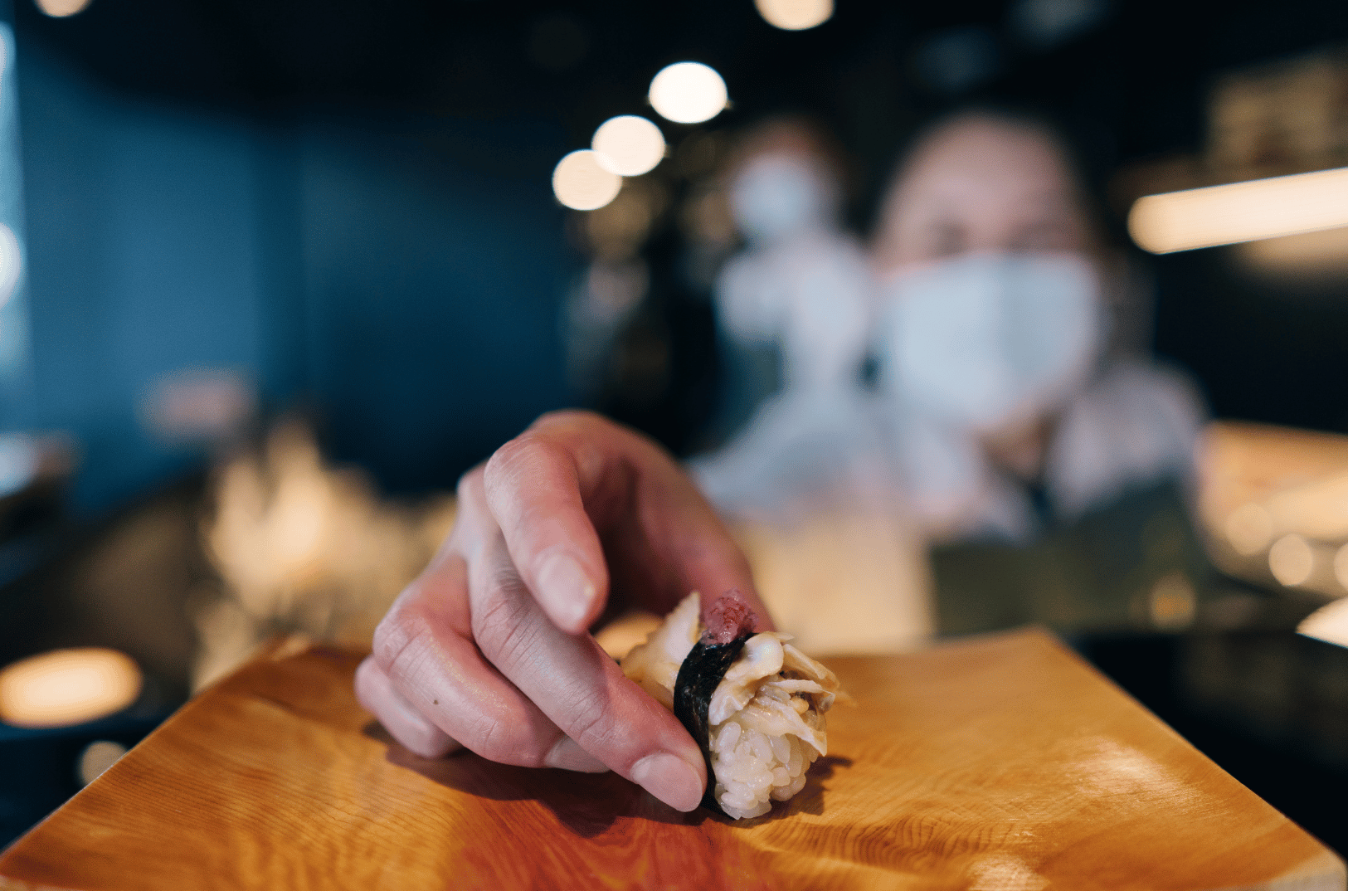 Close-up of a chef’s hand presenting a piece of sushi—wrapped in seaweed and topped with shellfish—on a wooden plate, set against a softly lit restaurant backdrop. A masked figure in the background and warm bokeh lighting evoke the intimacy and ceremony of omakase service. The gesture captures the final act of sourcing: the moment an ingredient, meticulously chosen and prepared, is offered with quiet reverence to the diner.