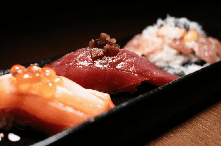 Close-up of three artisan sushi pieces—tuna, salmon, and seared nigiri—arranged on a black lacquer tray, highlighting precise knife work for an omakase feature.