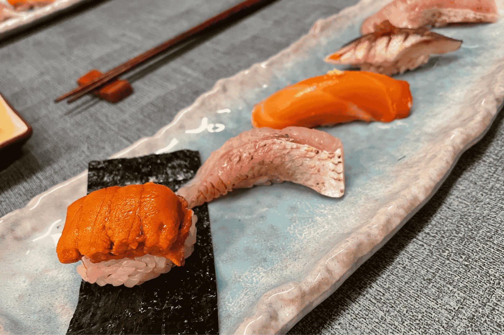 Close-up of an omakase plate with uni, salmon, and seared cuts of fish nigiri arranged in a row, highlighting knife work and presentation; chopsticks rest nearby on a textured table.