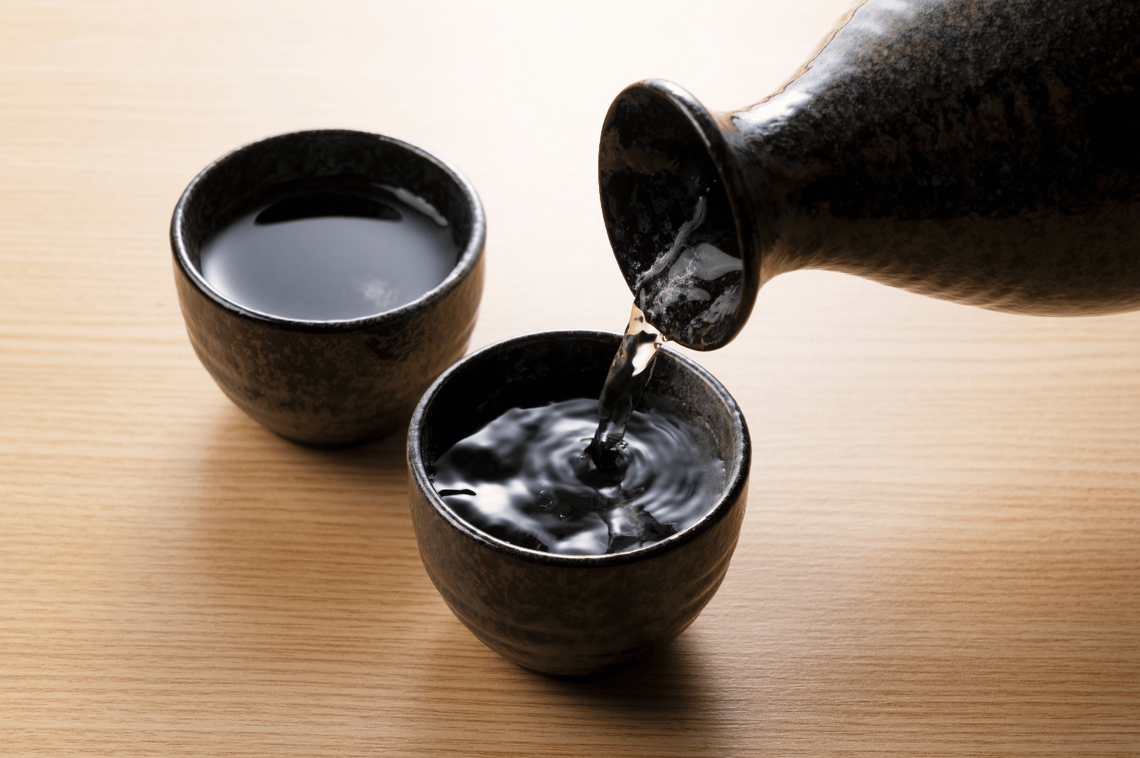 Rustic ceramic tokkuri pours dark premium sake into a textured ochoko on a pale wooden surface, ripples forming as the liquid meets the cup. The quiet tension between filled and filling vessels evokes the contemplative close of a refined Japanese meal. This intimate moment reflects Singapore’s elevated digestif programs, where post-celebration sake rituals extend the dining experience into a realm of sensory depth and cultural reverence.