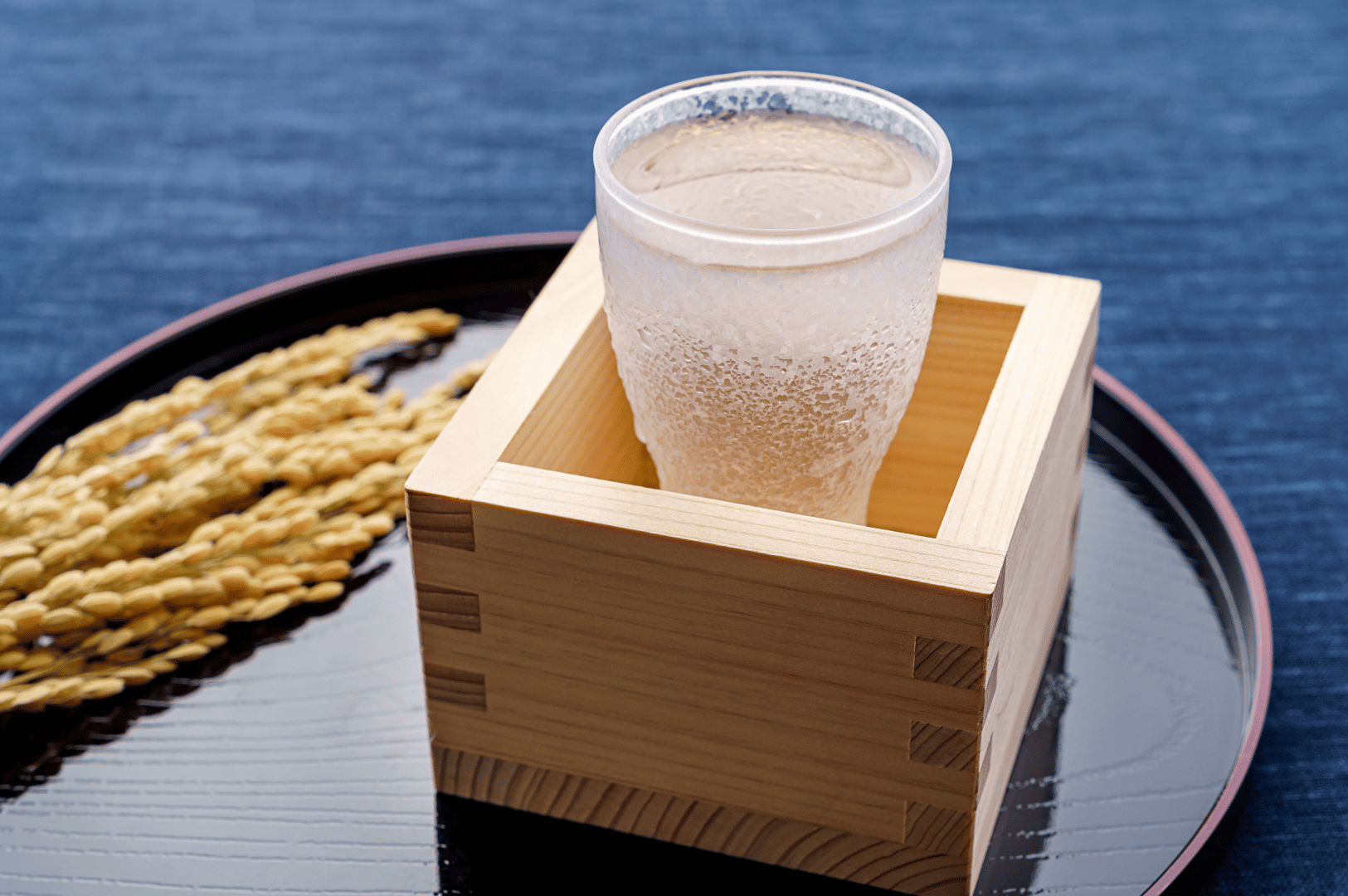 Chilled sake glistens inside a clear glass nestled in a traditional wooden masu, set atop a black lacquer tray with a red rim. A bundle of dried rice stalks rests beside the glass, symbolizing sake’s rice origins. The dark indigo backdrop and condensation on the glass evoke quiet luxury and reverence, reflecting Singapore’s refined digestif programs that extend Japanese culinary rituals into moments of post-celebration grace.