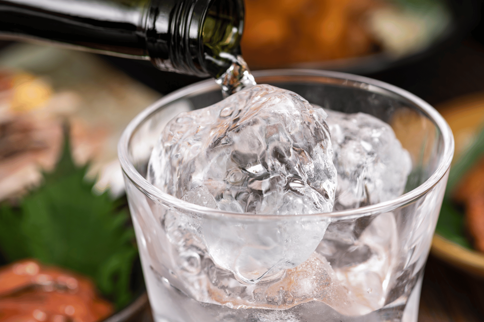Golden-hued liquid cascades into a clear glass filled with pristine ice cubes, the pour caught mid-motion against a softly blurred dining backdrop. The chilled clarity and dramatic contrast evoke a refined digestif ritual—where Singapore’s elevated sake and spirit programs extend Japanese cuisine into moments of quiet indulgence and sensory closure.