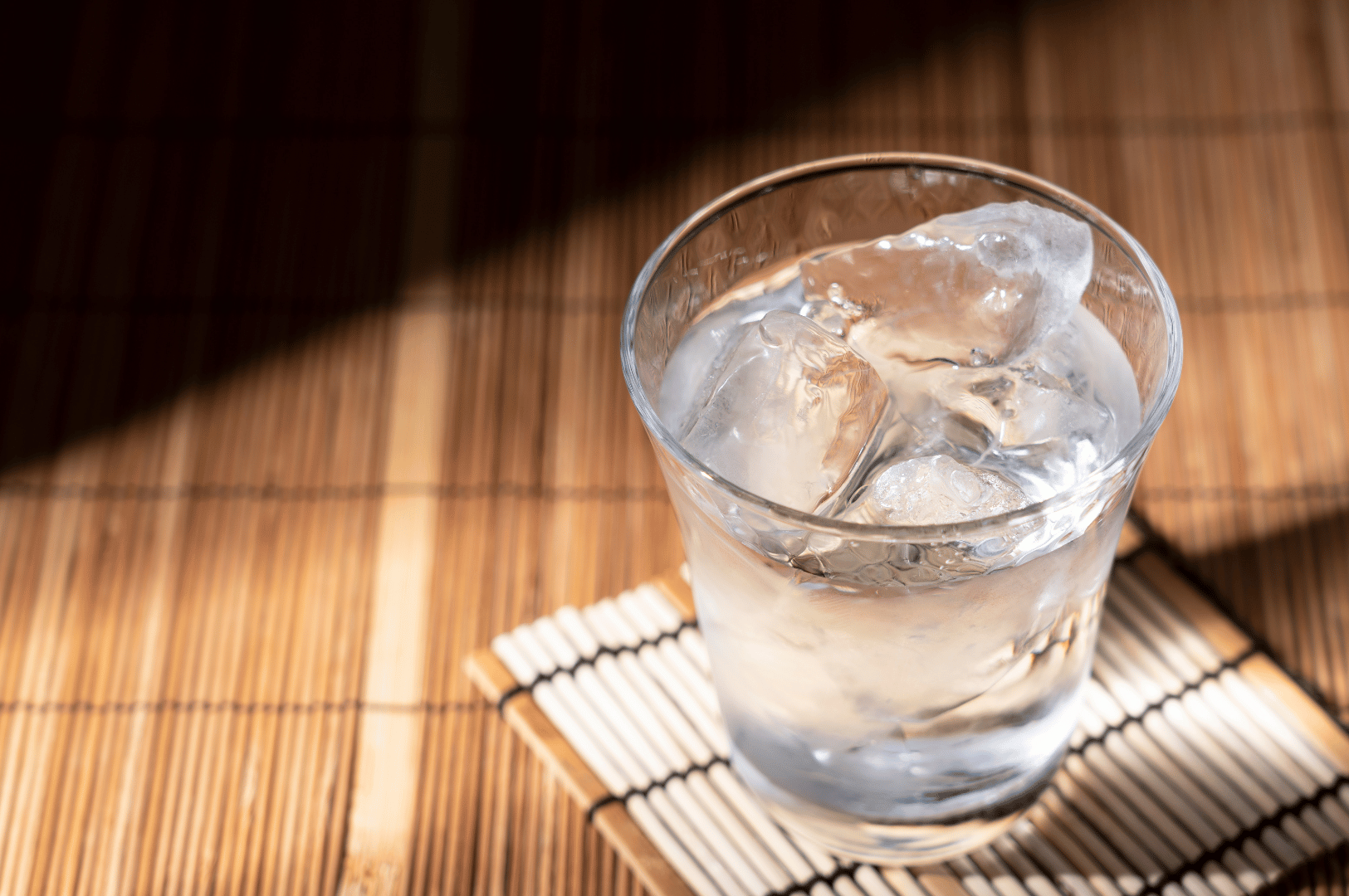 Clear glass of ice water rests on a sunlit bamboo mat, condensation glistening as shadows stretch across the surface. The partially melted ice and warm natural textures evoke a moment of calm and refreshment—an understated finale to Singapore’s elevated Japanese digestif programs, where even water becomes part of the sensory ritual that lingers after the last pour.
