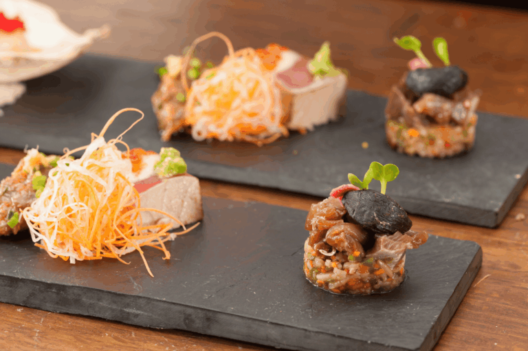 Three artfully plated servings of Singapore-Japanese fusion cuisine on black slate, featuring cylindrical tartare-style stacks topped with shiitake mushrooms, shredded local vegetables, and seared tuna slices beside avocado purée—highlighting the harmony of Japanese technique and Singaporean ingredients through vibrant textures and elegant composition.