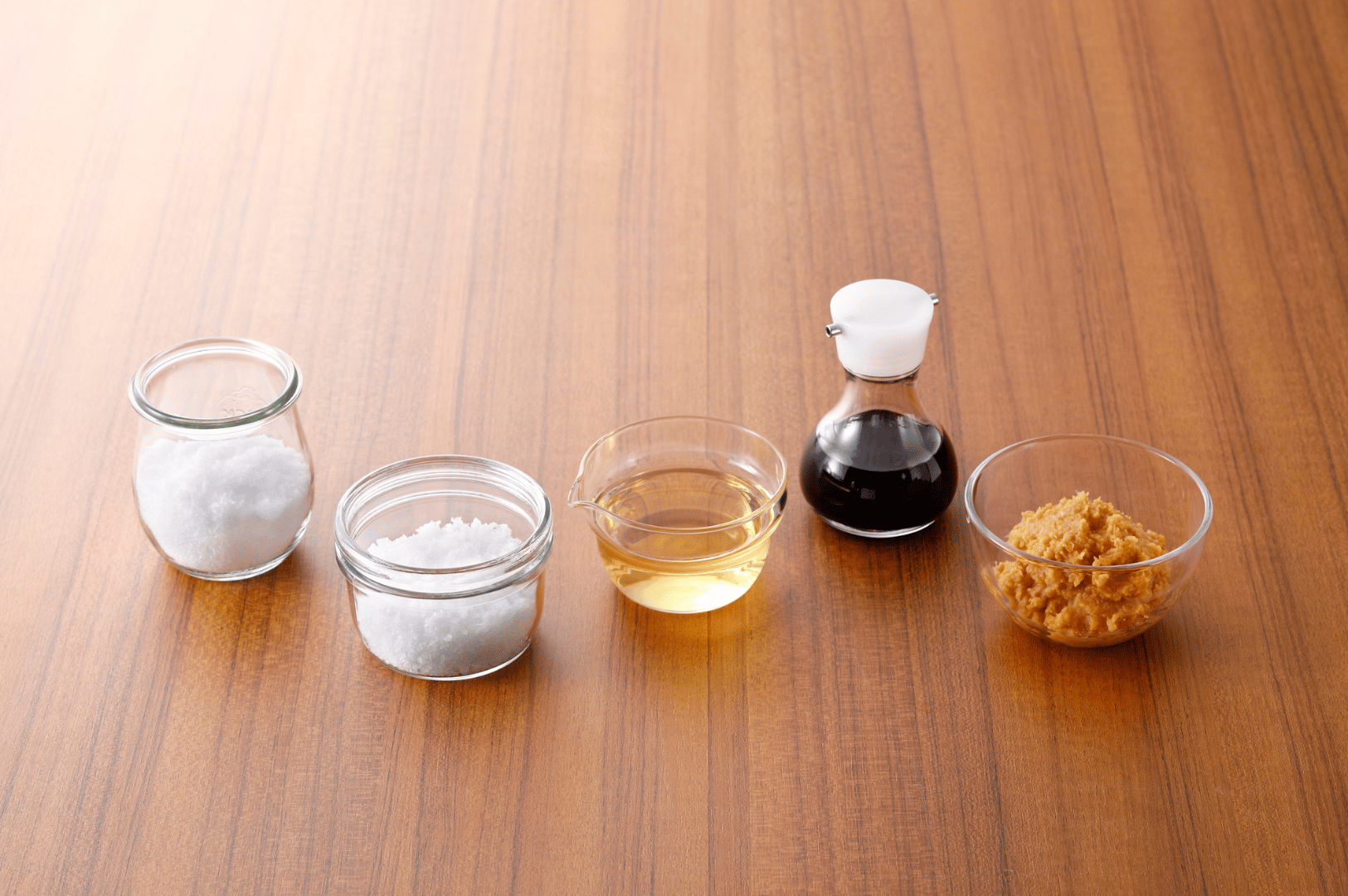 Five essential Japanese condiments—sugar, salt, rice vinegar, soy sauce, and miso paste—neatly arranged in ceramic and glass containers on a wooden surface, showcasing the foundational flavors that, when paired with Singaporean ingredients, elevate traditional Japanese cuisine into vibrant fusion.