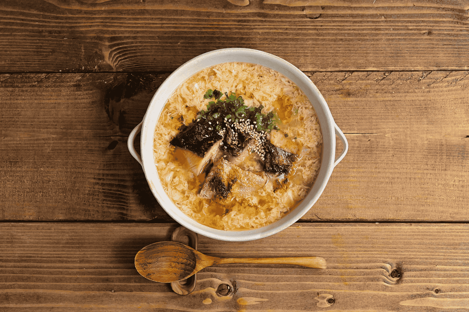A rustic bowl of Japanese-style egg drop soup garnished with seaweed, sesame seeds, green onions, and shredded meat, served with a wooden spoon on a natural wood surface—evoking comfort and tradition while inviting reinterpretation through Singapore’s bold, local flavors.