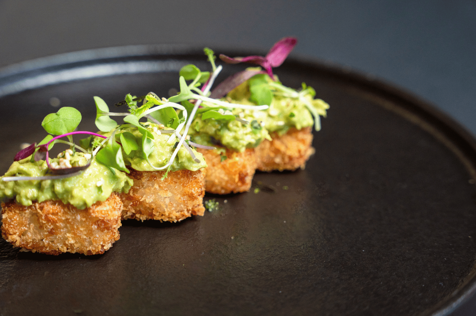 Close-up of a plated vegetarian appetizer from a Singapore omakase menu, featuring crispy golden croquettes or tofu topped with creamy avocado spread and garnished with microgreens and edible leaves. Presented on a dark ceramic plate, the dish balances rich textures and vibrant colors, reflecting the refinement and innovation of plant-based Japanese fine dining.