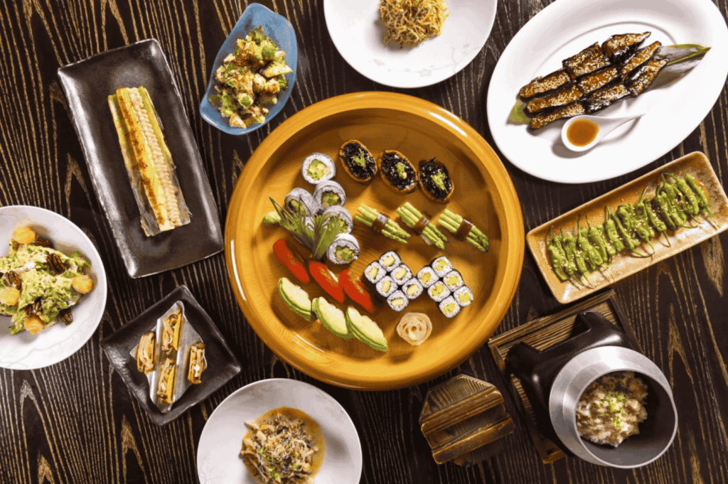 A vibrant vegetarian omakase feast arranged on a dark wooden table, showcasing a central wooden platter of sushi rolls made with seaweed, cucumber, and vegetable nigiri. Surrounding dishes include grilled plant-based skewers, tempura, rice bowls, and fresh salads, all meticulously plated in ceramic and metalware. The composition celebrates the diversity and artistry of plant-based Japanese cuisine in Singapore’s fine dining scene.