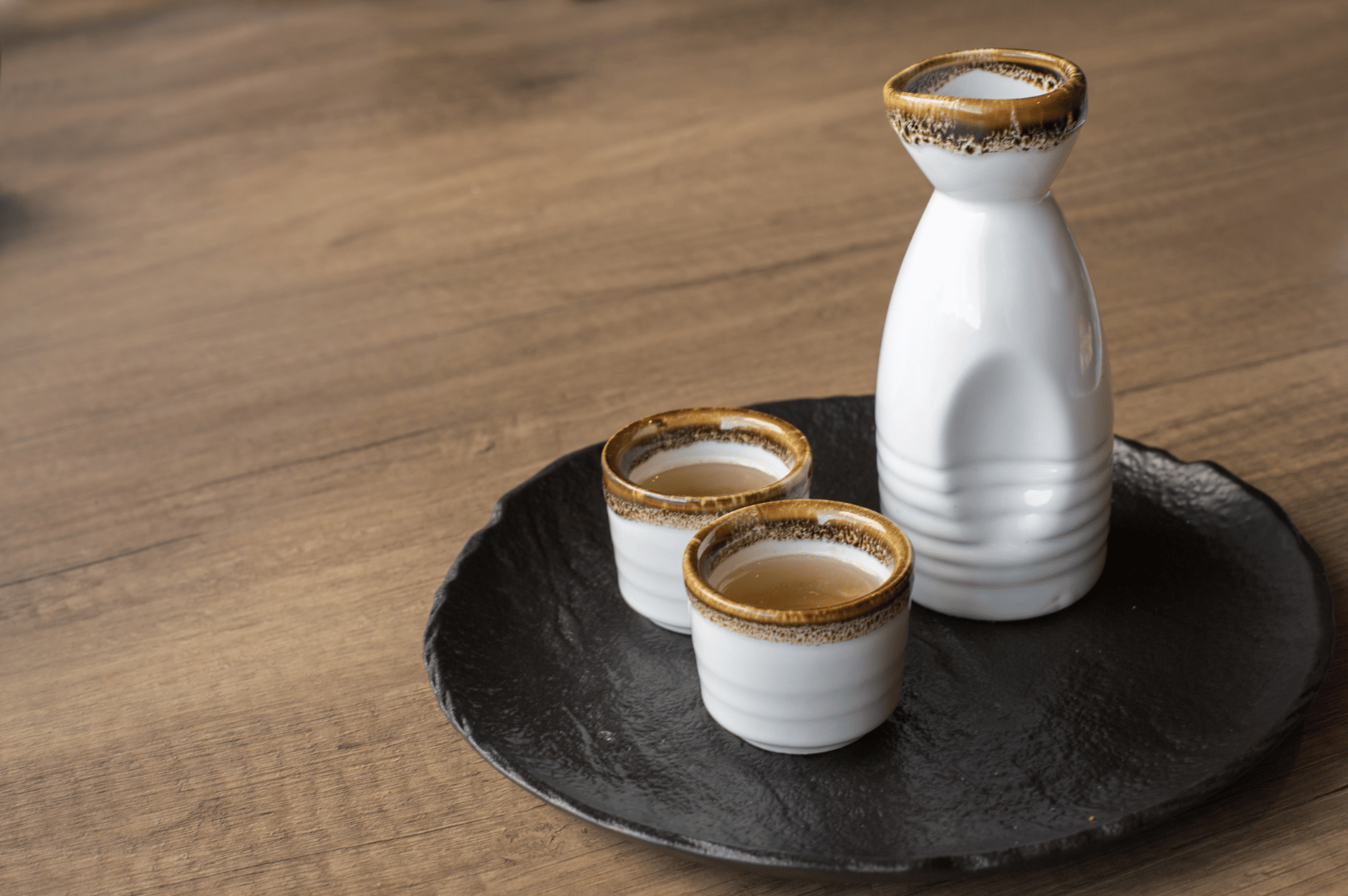 A handcrafted Japanese sake set on a dark round tray, featuring a white ceramic tokkuri and two ochoko cups filled with amber-hued sake. The rustic glaze and textured finish evoke traditional craftsmanship, while the serene presentation atop a wooden surface reflects the contemplative spirit of plant-based omakase dining in Singapore.
