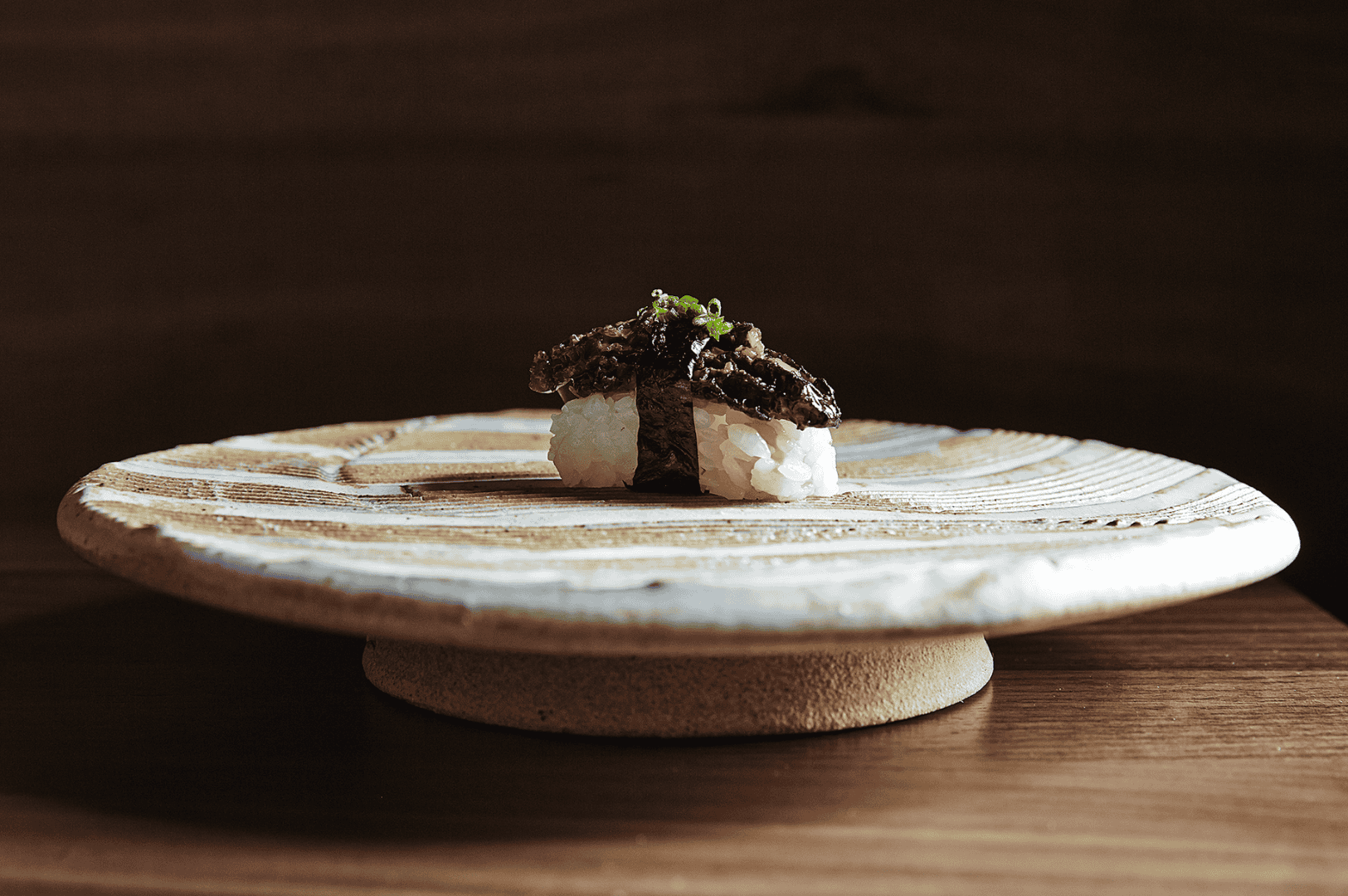 A single piece of vegetarian sushi elegantly plated on a textured ceramic dish with concentric grooves, featuring white rice topped with marinated mushroom and a sprinkle of scallions. The rustic plate and wooden backdrop evoke wabi-sabi aesthetics, emphasizing simplicity, craftsmanship, and the quiet sophistication of plant-based Japanese fine dining.