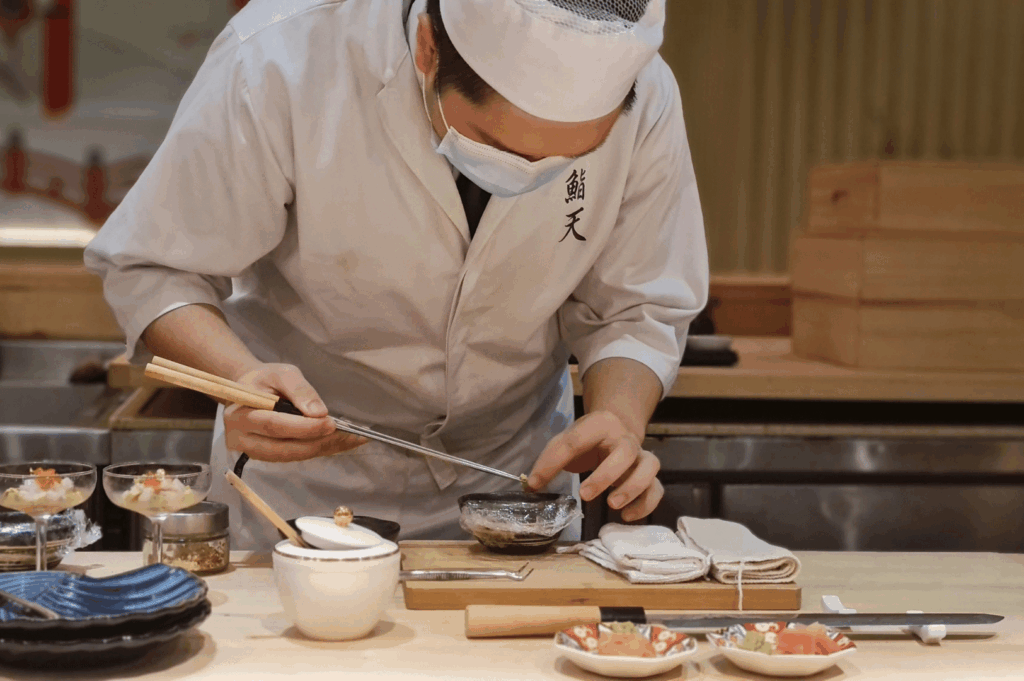 A masked chef at Ginza Sushi Ichi delicately plates a dish using chopsticks, surrounded by refined mise en place—seasoning bowls, folded cloths, and layered seafood glasses. The crisp white uniform and focused gesture evoke precision and performance, reinforcing the restaurant’s theatrical approach to omakase and its elevated value proposition in contrast to Shinji’s quiet restraint.