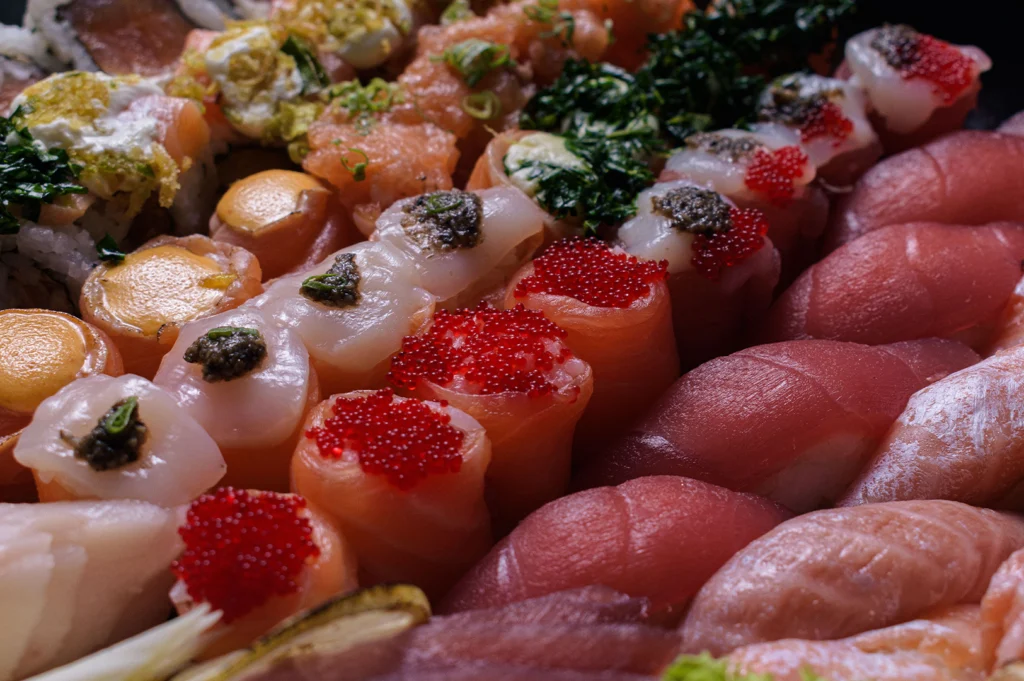 A vibrant close-up macro shot displays a dense, luxurious array of fresh sushi and nigiri tightly arranged together. The selection showcases salmon nigiri generously topped with bright red tobiko (flying fish roe), delicate scallops garnished with dark caviar, tuna nigiri, rolled sushi lined with chopped herbs, and creamy, sauce-drizzled pieces.   
