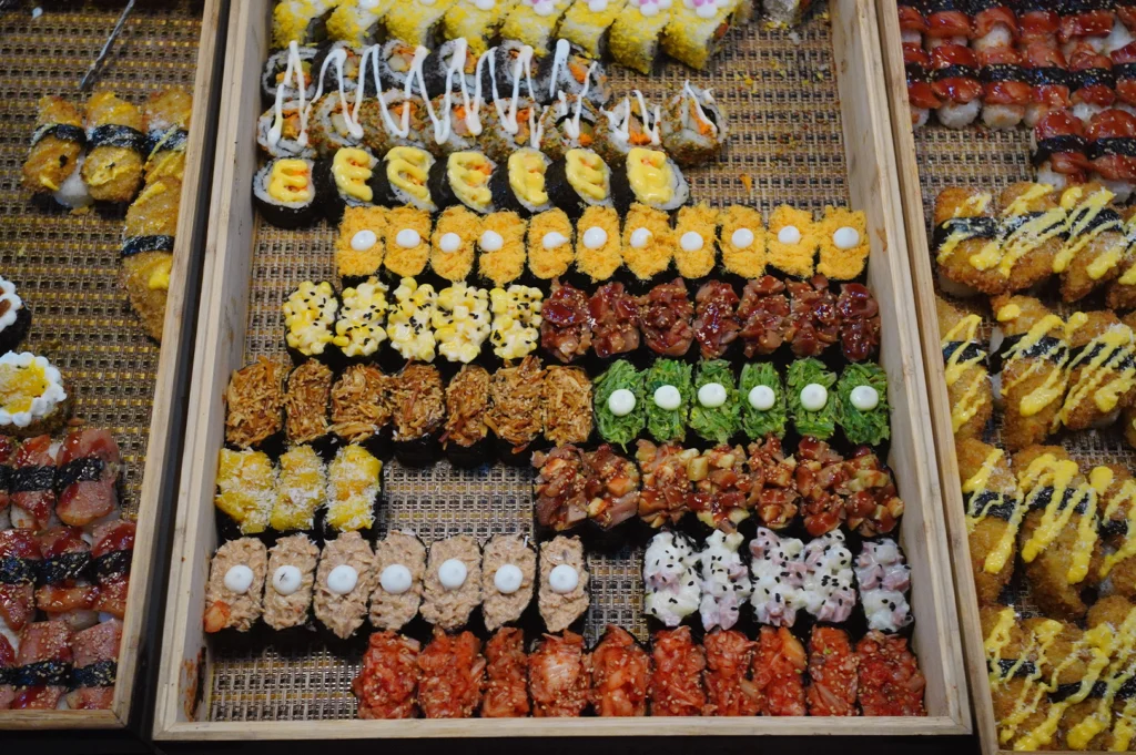 A high-angle overhead view shows a large wooden buffet tray neatly packed with neat, colorful rows of diverse maki rolls and gunkan sushi. The elaborate spread includes rolls coated in yellow tobiko, seaweed-wrapped pieces topped with mayo-drizzled corn, bright green wakame seaweed salad, seasoned minced tuna, red kimchi, potato salad, and crispy deep-fried appetizers lined up along the sides.   
