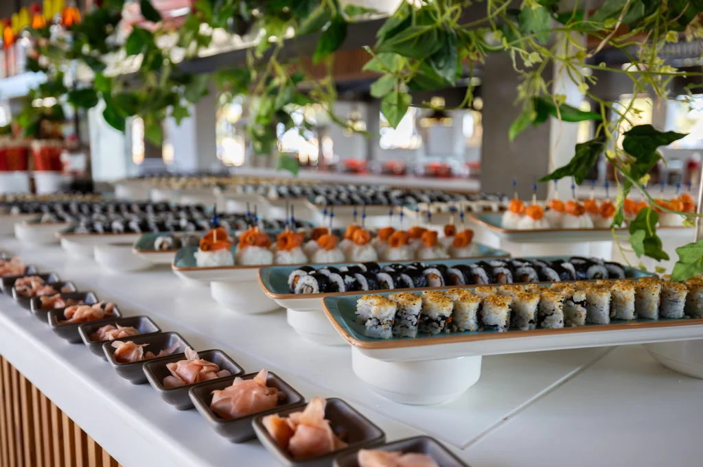 A wide perspective shot captures an expansive hotel buffet counter beautifully arranged with long ceramic platters of freshly made sushi rolls and nigiri. Lush green faux vines hang gracefully overhead while neat rows of small, square dishes containing pickled ginger line the foreground, leading toward tiers of sesame-crusted rolls and rice bites topped with savory toppings.   
