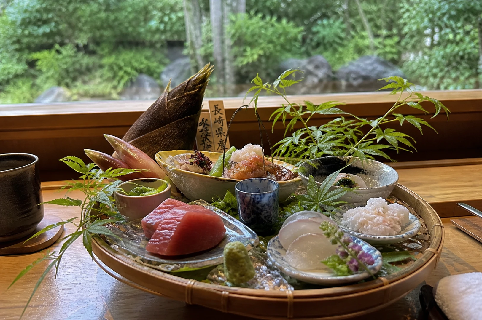 An elaborate and artfully arranged Japanese Kaiseki sashimi platter is presented in a circular bamboo basket, set against a serene backdrop of a lush green garden visible through a large window. The assortment features thick slices of maguro (tuna), delicate white fish, and meticulously prepared seafood served in various small ceramic bowls. A large, dark brown bamboo shoot and pink ginger buds provide striking vertical elements, while fresh green maple leaves and a purple flower sprig add organic beauty to the arrangement. A small wooden plaque with Japanese calligraphy stands within the basket, and a dark ceramic teacup sits on the wooden table to the left, completing this high-end traditional dining scene.