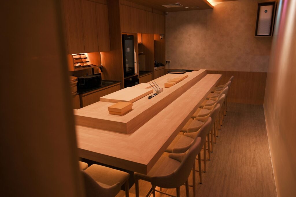 A high-angle view of a minimalist Japanese omakase sushi counter featuring light wood grain finishes, warm ambient lighting, and sleek modern chairs in an intimate dining room setting.