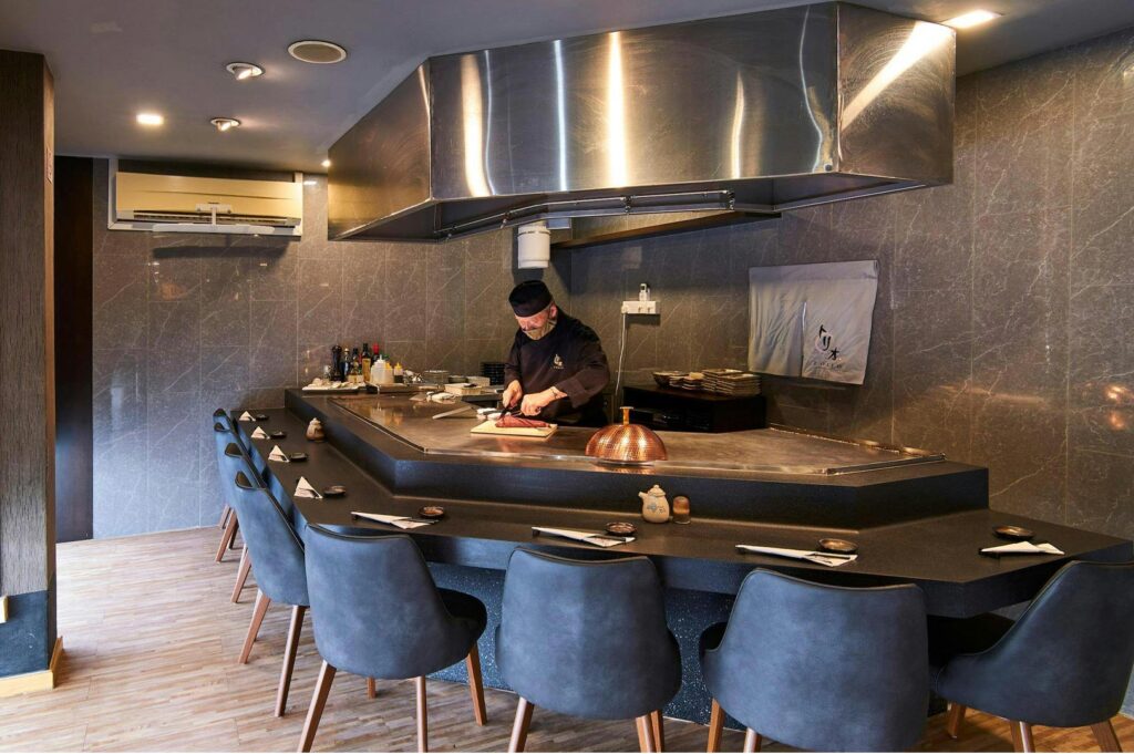 A professional teppanyaki chef in a black uniform and face mask slicing a steak on a large stainless steel flat-top grill in a modern restaurant with gray marble walls.