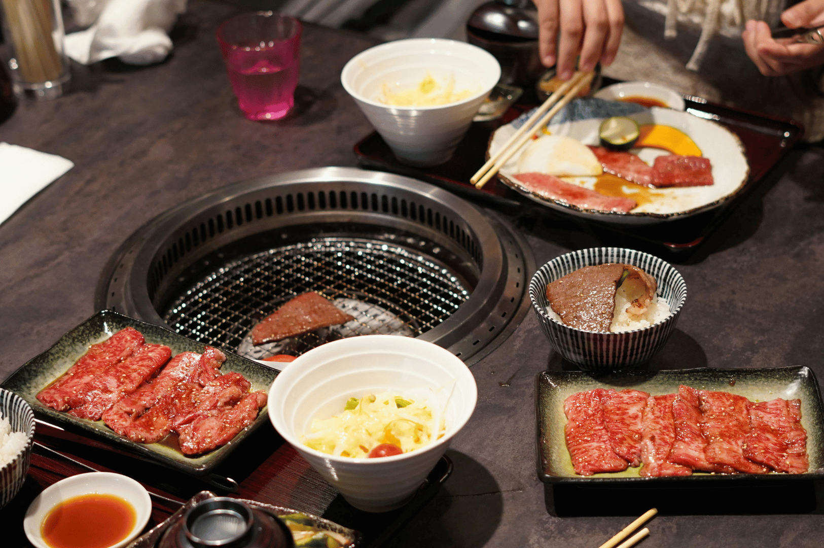 People grilling sliced beef and vegetables at a Japanese BBQ table, with small bowls and chopsticks