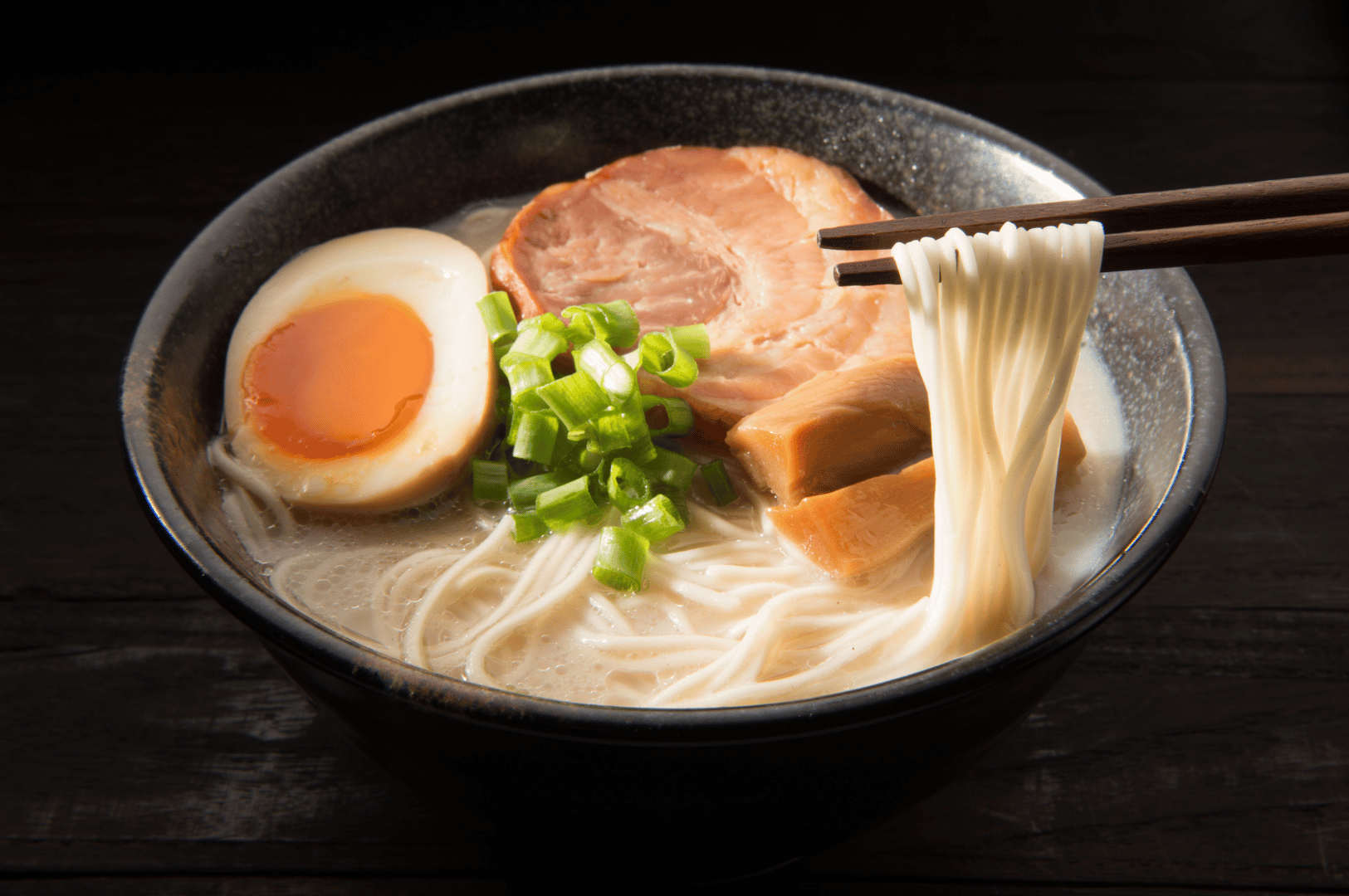Close-up of a steaming bowl of ramen with sliced pork, soft-boiled egg, and green onions—noodles lifted by chopsticks.