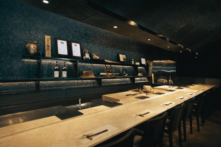 Warm, dimly lit omakase counter at Kuriya Dining with neatly set seats, wooden boards, and a display of sake and chef awards on dark textured shelves.