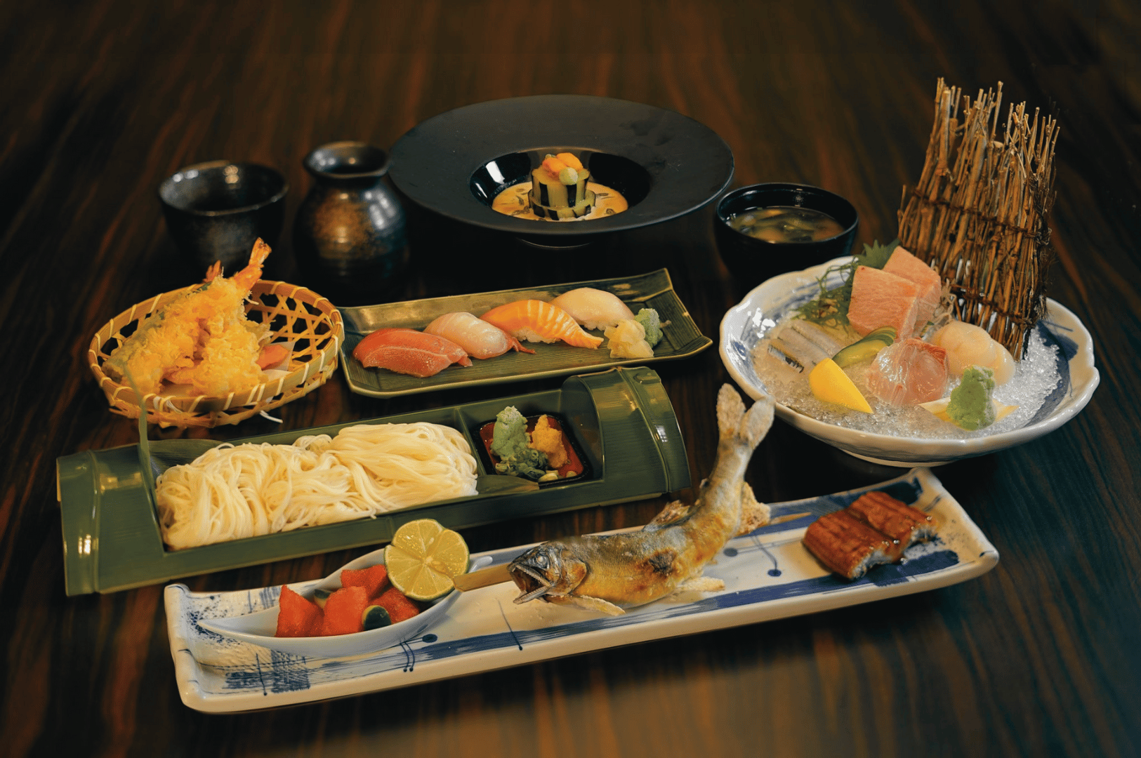 A curated omakase spread at Kuriya Dining featuring sushi, sashimi on ice, grilled fish, tempura, cold noodles, and small seasonal sides arranged on a wooden table.