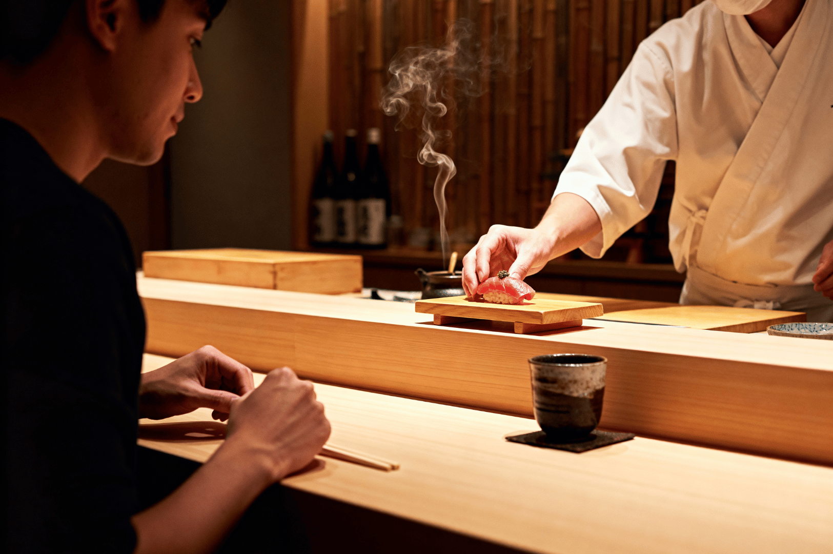 A sushi chef in a white uniform serves freshly prepared nigiri on a wooden board to a guest at a traditional sushi counter, capturing the intimate chef’s table omakase experience in Singapore.