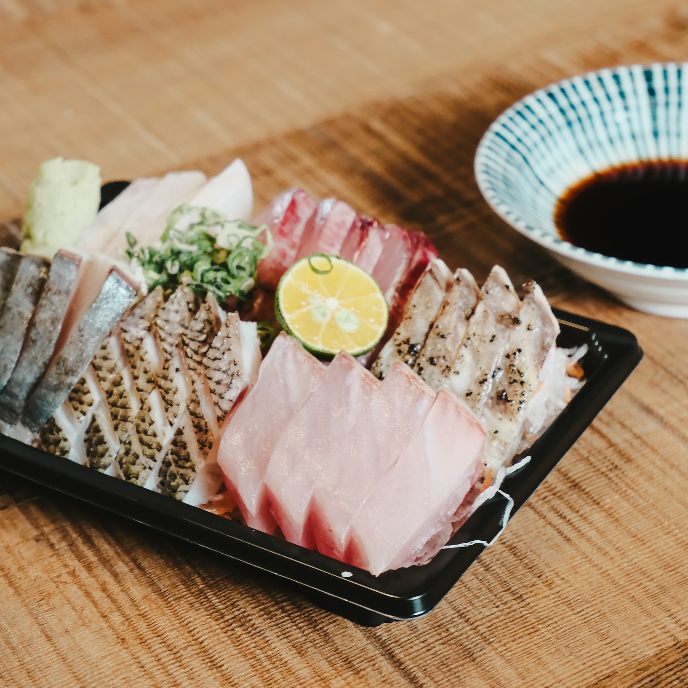An assorted sashimi platter with different types of fish, lime, and wasabi, beside a soy sauce bowl.