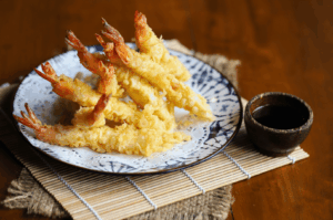Golden shrimp tempura arranged in a precise fan on a blue-and-white ceramic plate, served with a dark tentsuyu dipping sauce on a woven mat—capturing the meticulous presentation and crisp texture central to Yamato’s tempura omakase in Singapore, as examined through expert culinary critique.
