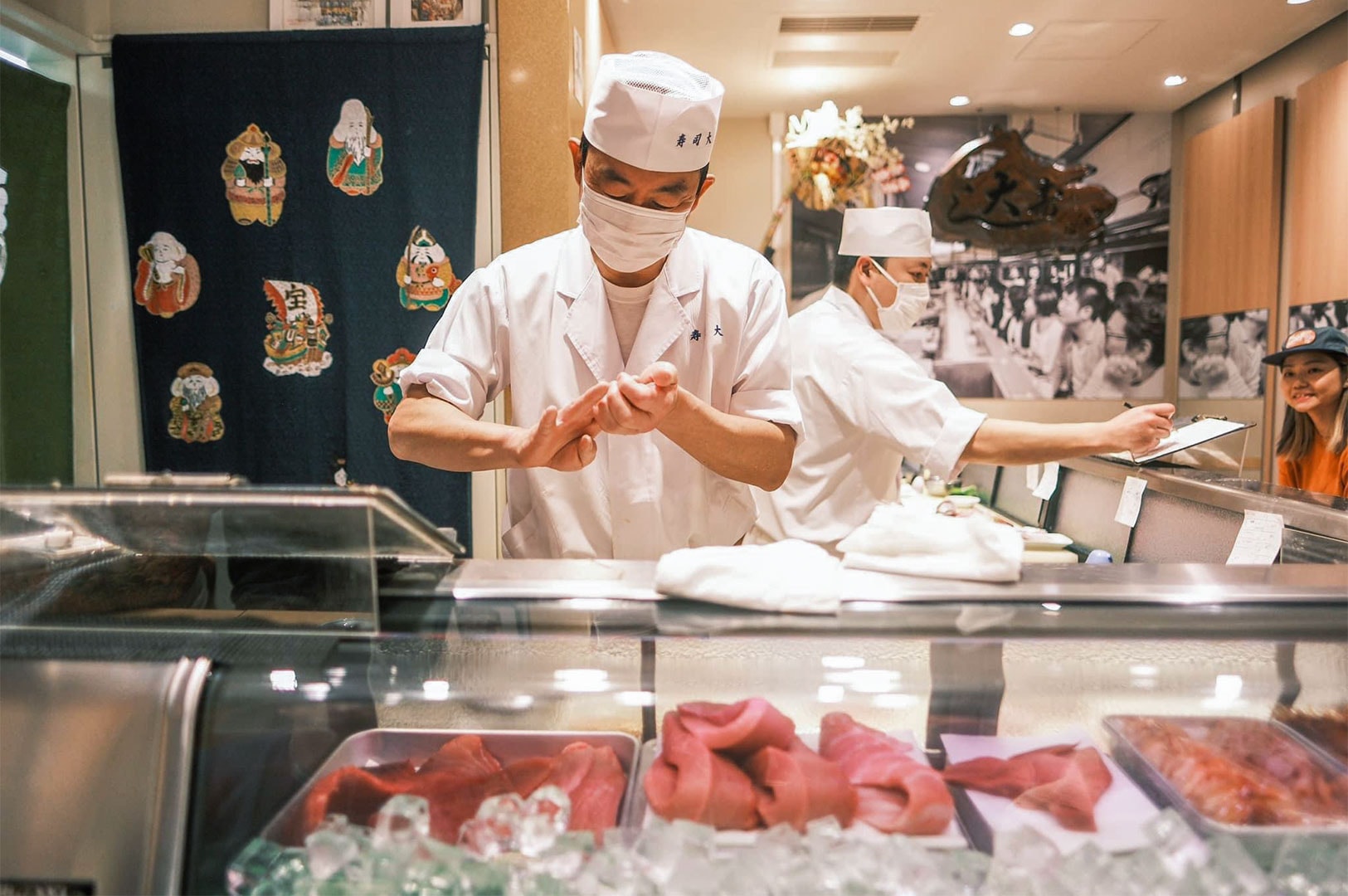 A sushi chef in a white uniform and face mask shaping sushi rice behind a glass display case filled with fresh tuna cuts.